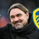 Leeds United manager Daniel Farke holding a thumbs up and smiling next to a Leeds badge.